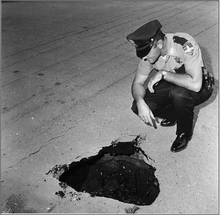 Photograph of Police Officer looking at pothole in roadway.